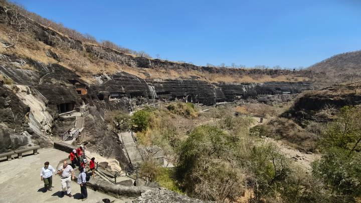 Panoramic view across an arid valley revealing a row of rock-cut Ajanta caves in a cliffside, with tourists on a walkway.