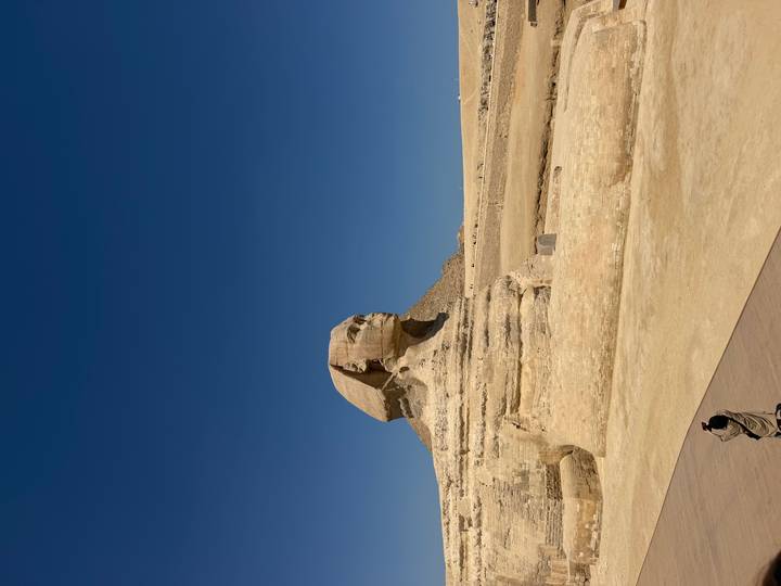 The Great Sphinx of Giza under a cloudless deep blue sky with a tourist photographing in the foreground.