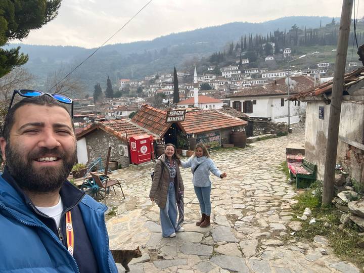 Smiling travelers pose on a cobblestone lane overlooking a hillside village of white houses and tiled roofs.