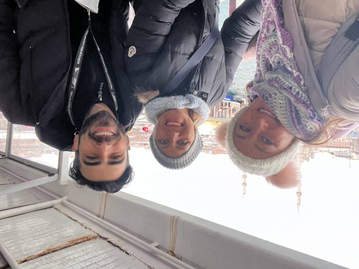 Three friends bundled in winter gear smile on a Bosphorus ferry with Istanbul mosques behind.