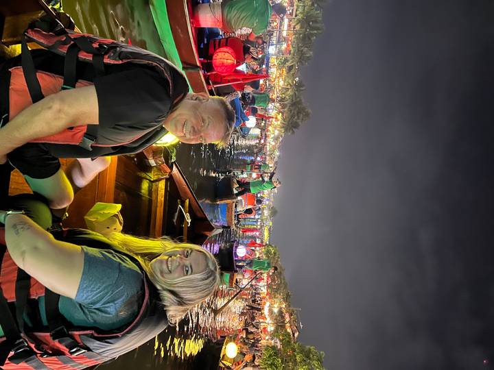 Two tourists seated in a lantern-lit boat among many colorful boats on a night river festival