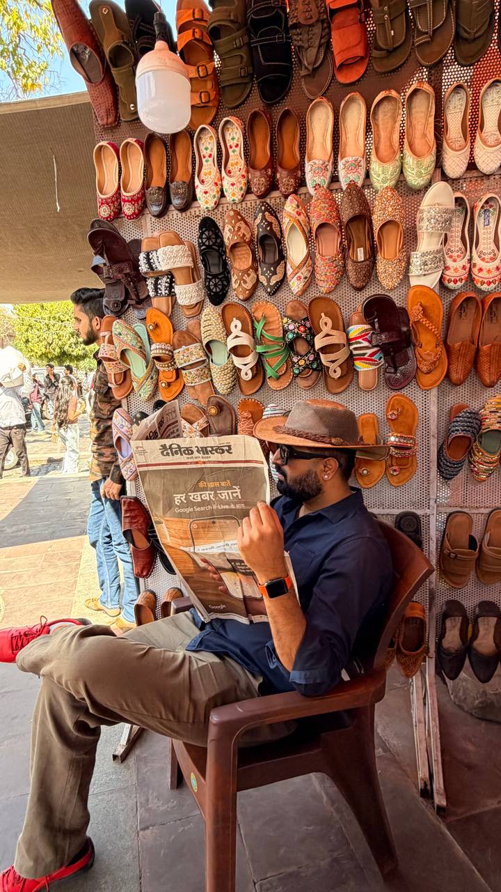 Man reading a Hindi newspaper at a vibrant street stall selling colourful sandals in India.