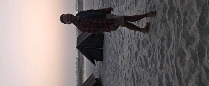 Man walking near tents on a dimly lit sandy riverside camp at dusk.