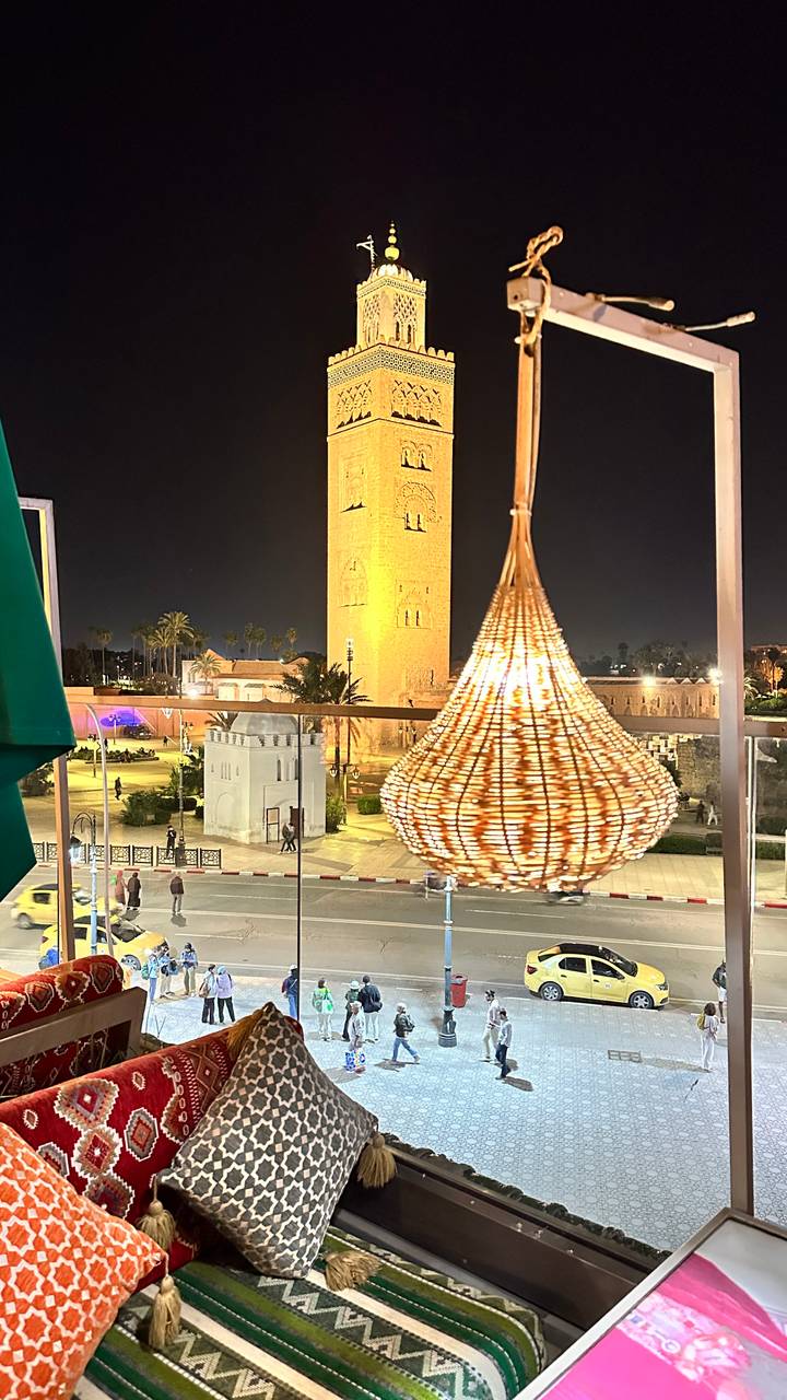 Night view of Koutoubia Mosque tower in Marrakesh seen from a rooftop bar with a woven lantern in the foreground.