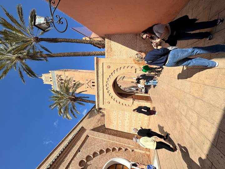 Visitors gather outside the brick gateway of Koutoubia Mosque with tall minaret and palm trees against deep blue sky.