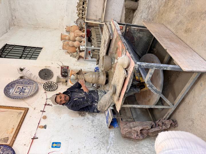 Potter shaping clay on a wheel inside a ceramics workshop surrounded by finished pots.