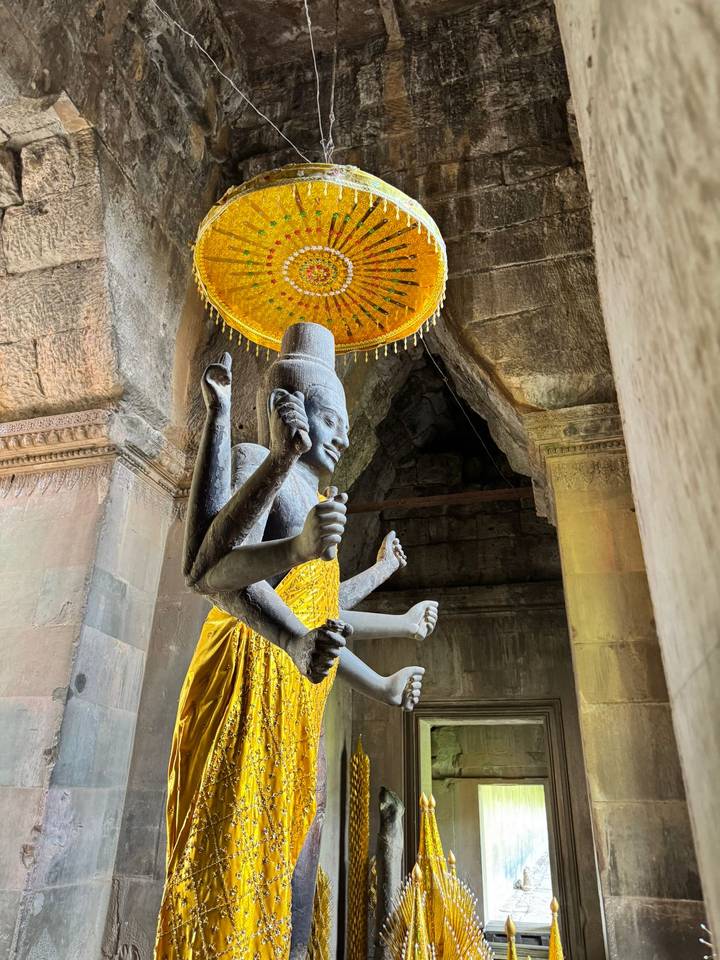 Multi-armed stone deity statue draped in a yellow cloth beneath a decorative parasol inside a temple corridor.