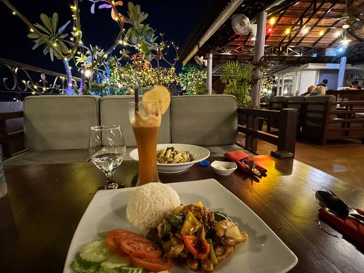 Outdoor evening dining table with rice, stir-fry dish and a tall fruit drink under decorative lights.