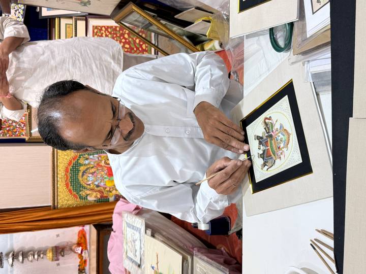 An artist in traditional attire meticulously painting a miniature artwork at a table.