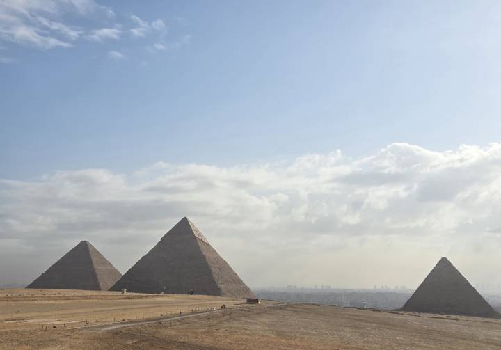 Panoramic view of three pyramids rising from the desert with a hazy city skyline beyond.