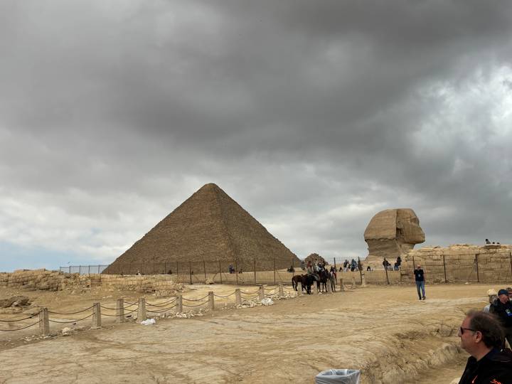 Wide scene of the Pyramid of Khafre and the Sphinx under a brooding cloudy sky with tourists nearby.