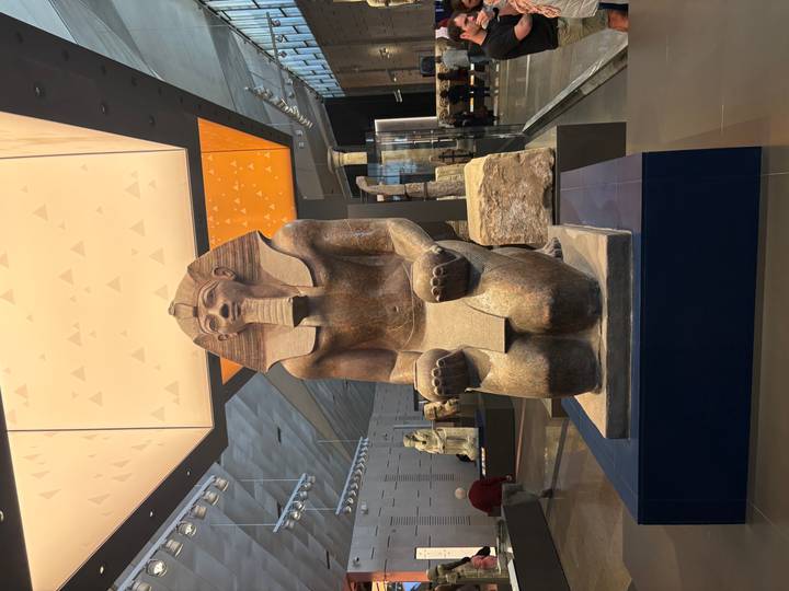 Large kneeling pharaoh statue displayed in a modern museum hall with geometric ceiling panels.