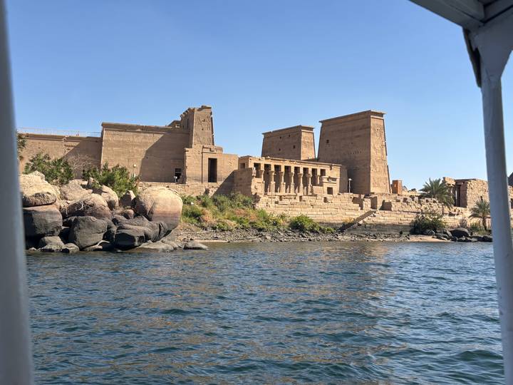View of Philae Temple complex on an island taken from a boat over rippling blue water.