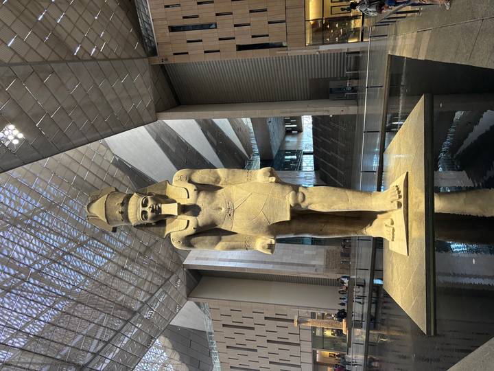 Colossal standing statue reflected in a shallow pool inside a vast modern museum atrium.