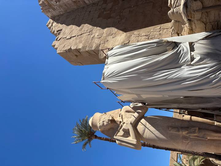 Large statue at Karnak Temple partially covered by protective sheets under a clear blue sky.