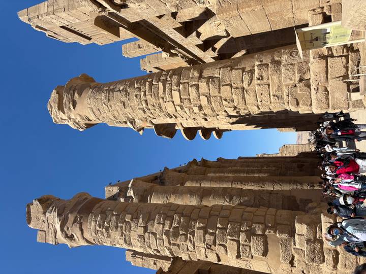 Crowds walk between towering sandstone columns of Karnak Temple under bright sunshine.