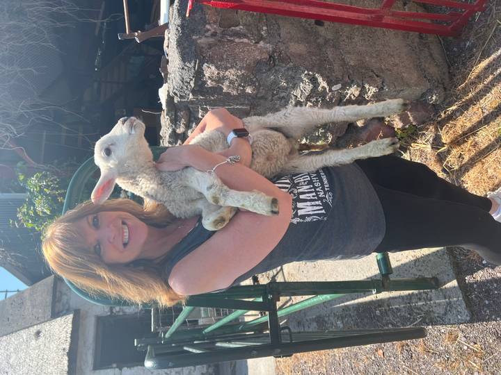 Smiling visitor holds a young lamb outside a rustic Irish farm building.