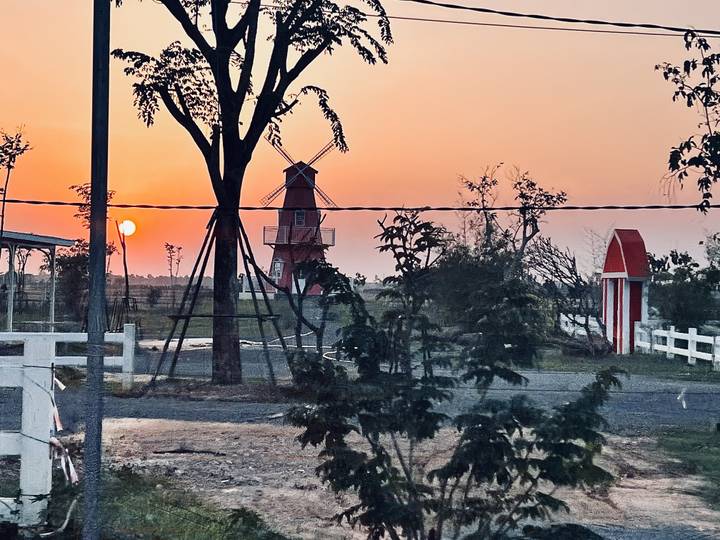 Rural sunset scene with orange sun setting behind a small windmill and scattered trees.