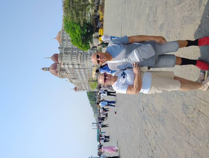Travellers posing in front of the historic Taj Mahal Palace Hotel and Gateway of India square under a clear blue sky in Mumbai.