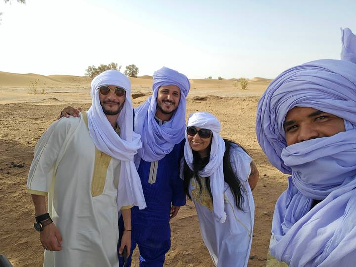 Four friends in traditional turbans smile amid rolling Sahara desert dunes.