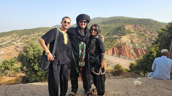 Three travelers in black outfits pose on viewpoint overlooking red and green Atlas foothills.