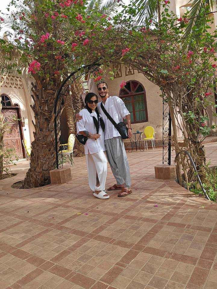 Couple in breezy white outfits embrace in shaded courtyard of Moroccan riad.