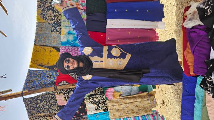 Man in blue robe spreads arms while surrounded by colourful fabric stalls at desert market.