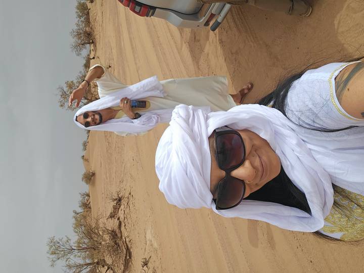 Smiling couple take selfie on sandy desert plain wearing white turbans and sunglasses.