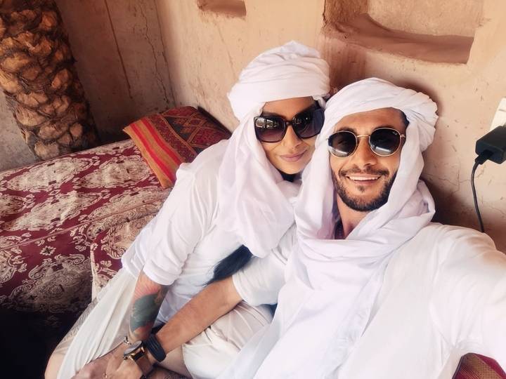 Couple relax on patterned cushions wearing white turbans and sunglasses in desert lodge.