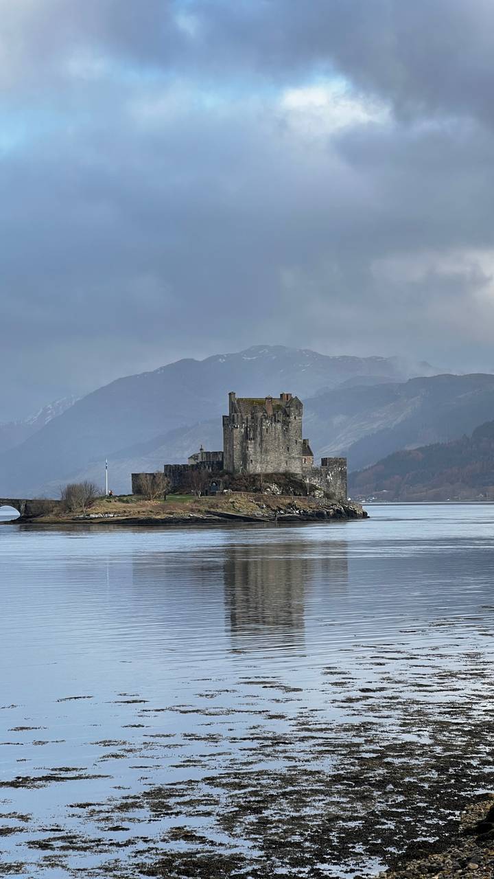 Iconic Eilean Donan Castle stands on a small island surrounded by still water against misty Highland mountains.