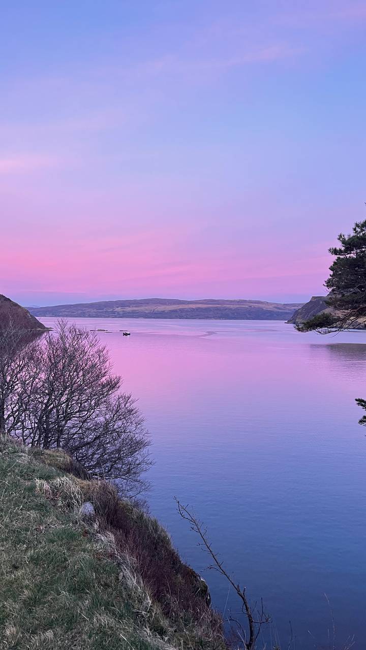 A tranquil loch reflects pastel pink and purple twilight hues with silhouetted hills on the horizon.