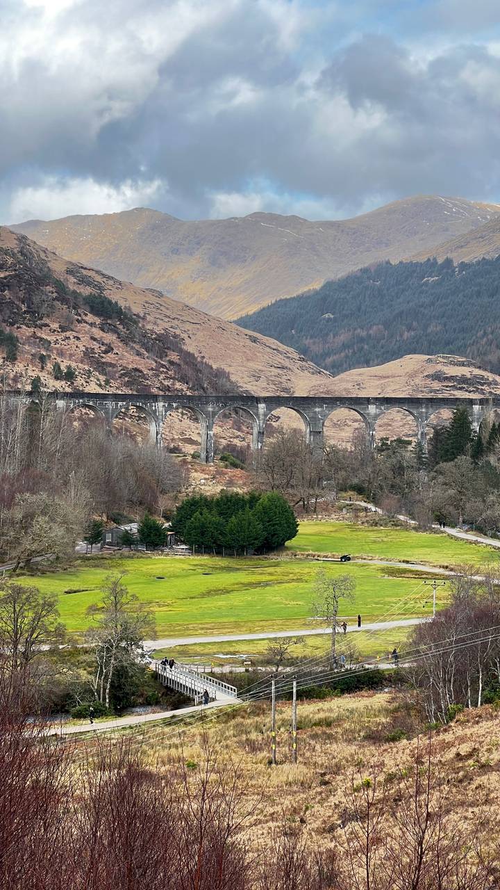 The curved arches of the Glenfinnan Viaduct span a wooded valley with rugged Highland hills behind.