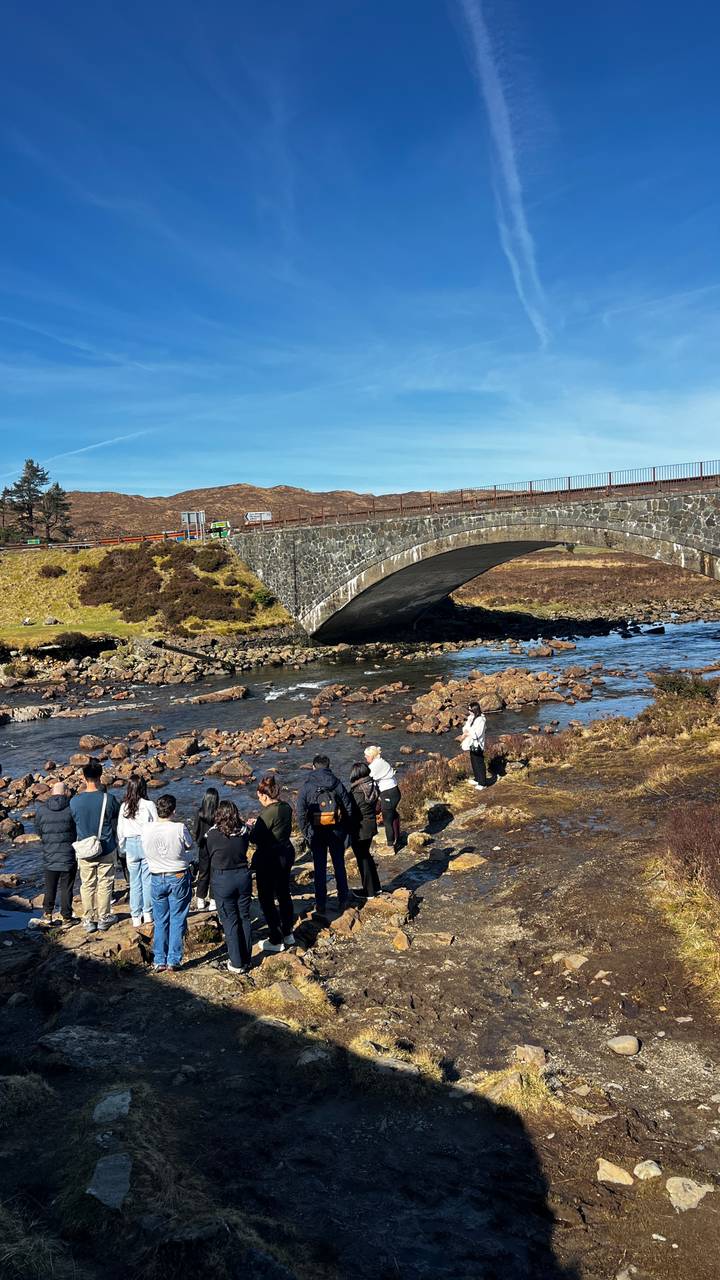 Tourists gather near an old stone arch bridge spanning a rocky Highland stream under bright sun.