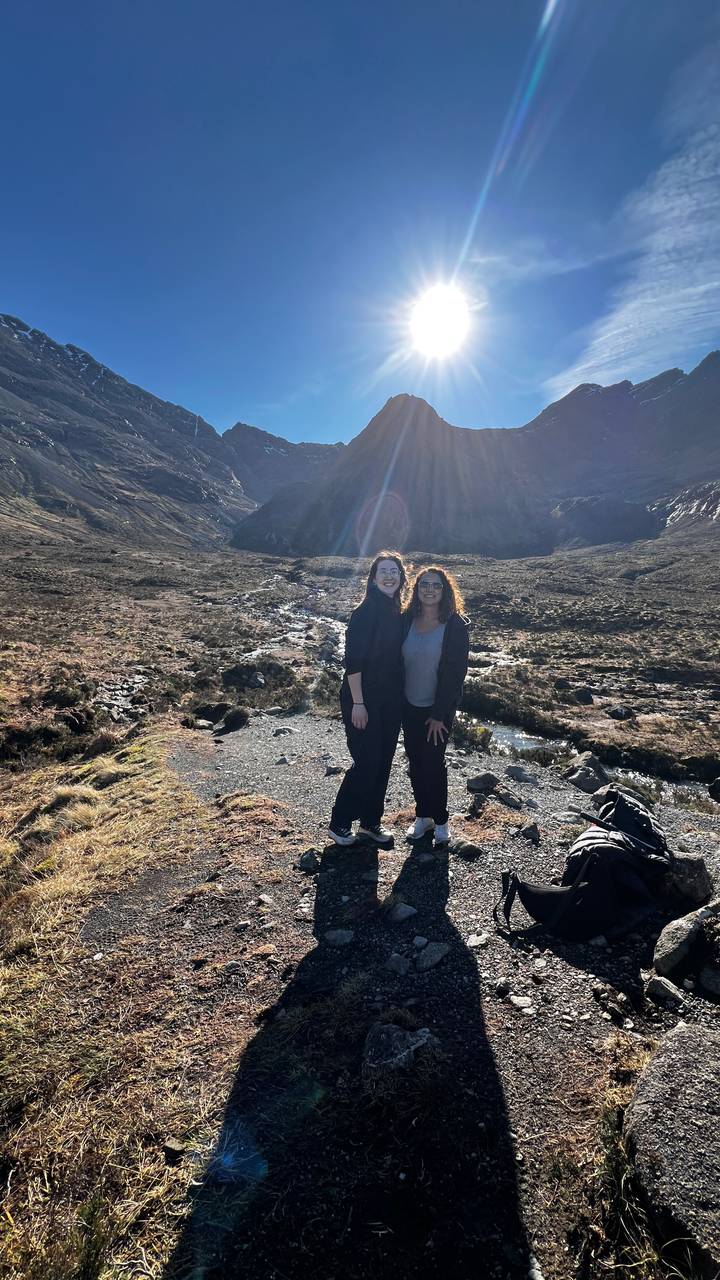 Two hikers smile for a photo in a rocky mountain valley with sunbeams shining over the peaks behind them.