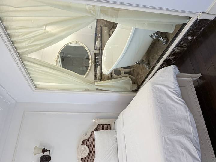 Elegant hotel room with white bed adjacent to bathroom featuring clawfoot tub behind glass wall