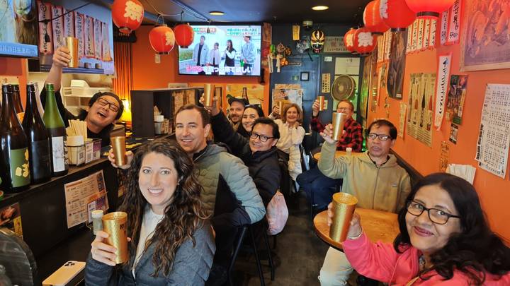 Happy travelers raise cups inside a cozy Japanese izakaya decorated with lanterns and posters.