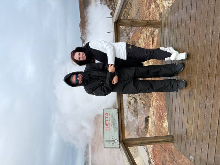 Bundled travellers pose on a wooden platform amid billowing geothermal steam and warning signs.