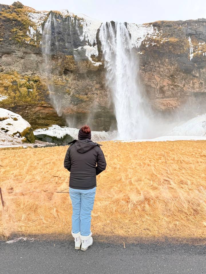 Person in winter clothing admires the plume of Seljalandsfoss waterfall crashing behind icy rocks.