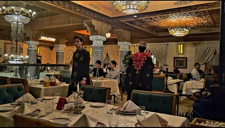 Dimly lit Moroccan restaurant with ornate décor, musicians and diners at candle-lit tables.