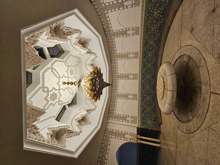 Ornate Moroccan interior with carved stucco ceiling, star motifs, large chandelier and circular stone fountain basin.
