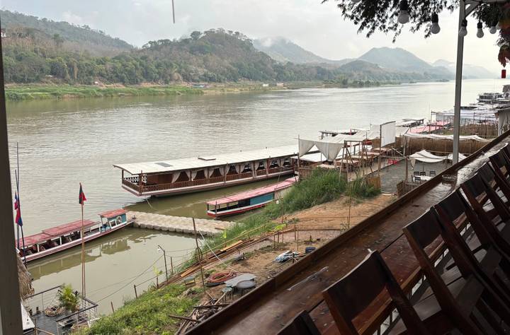 Long wooden boats moored along the Mekong River with forested hills beyond.