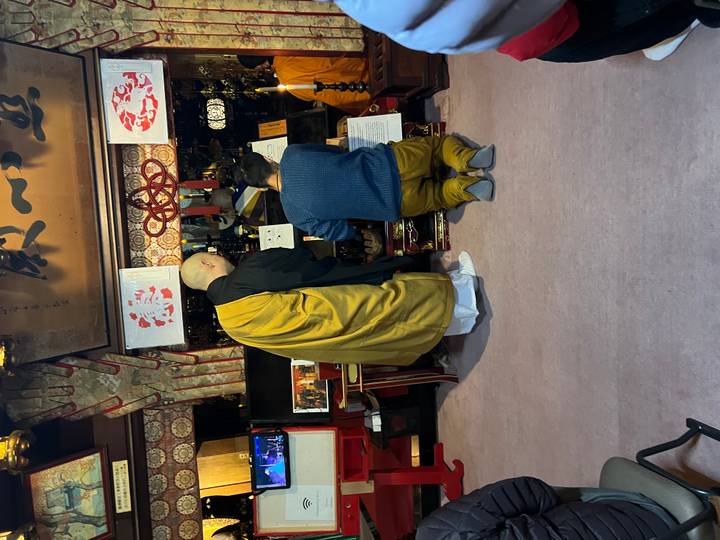 Buddhist monk instructs a visitor kneeling before a low altar in a dimly lit temple.