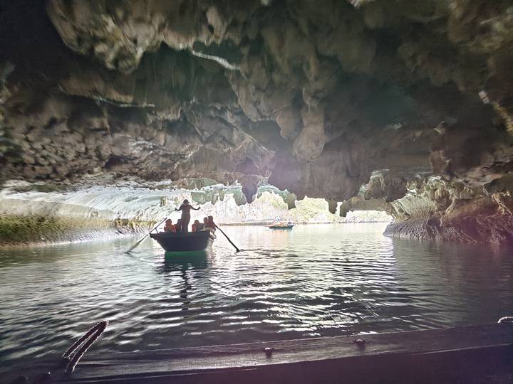 Small rowboat with passengers paddling through a limestone cave opening onto calm green water.