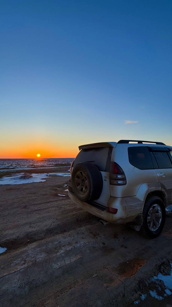 An off-road vehicle is parked on a barren, snowy desert plain under a glowing orange sunset.
