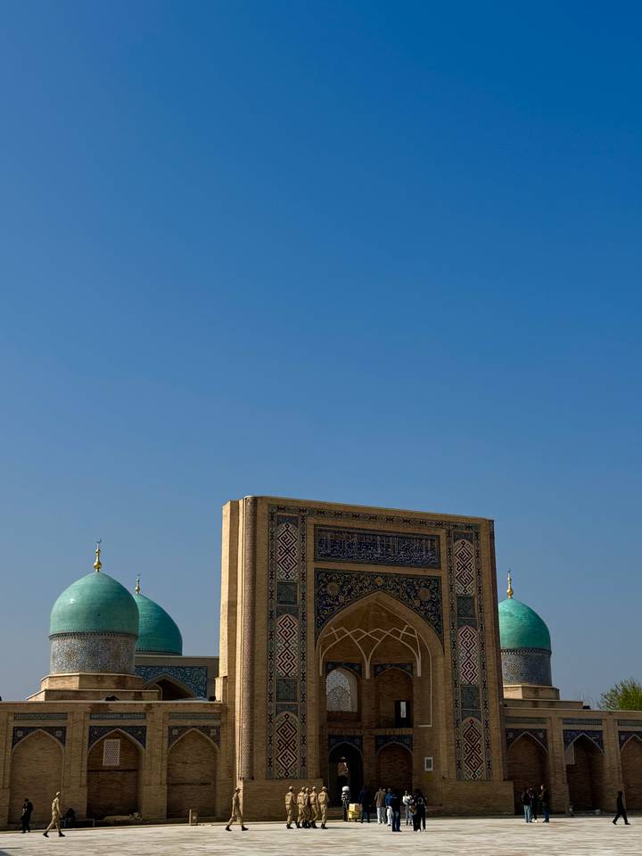 Blue domes and an ornate portal of a historic Islamic complex stand beneath a vast clear sky.