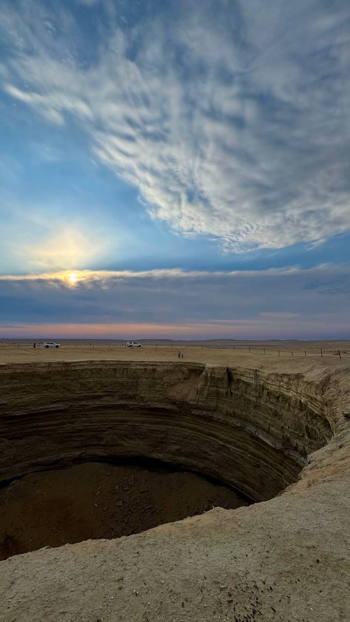 Cars and tiny figures stand at the rim of a massive desert sinkhole under a dramatic sky.