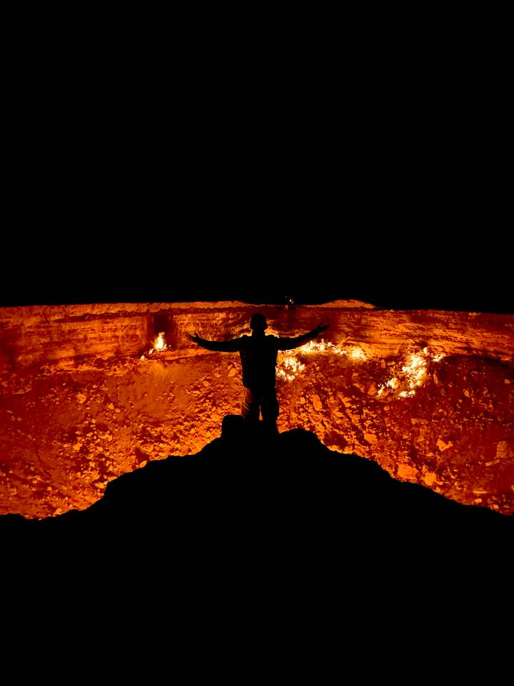 A silhouetted figure stands with arms outstretched before the fiery glow of the Darvaza gas crater at night.