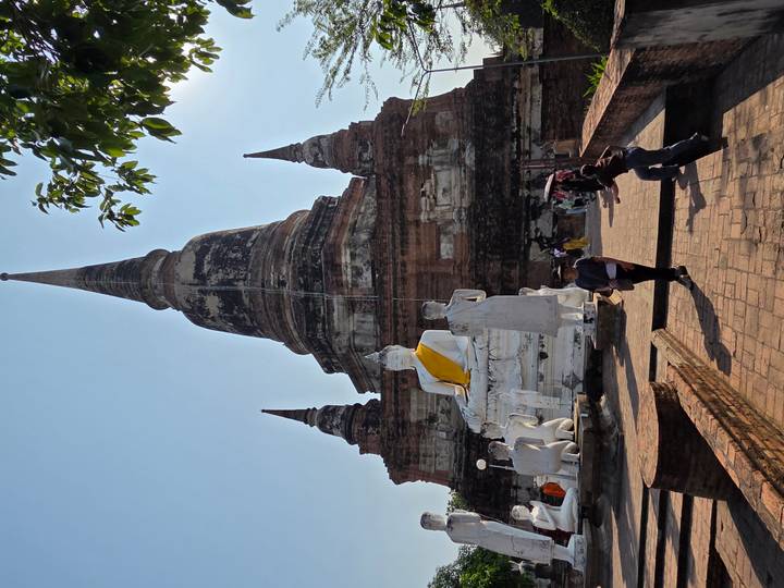 Tall ancient brick stupa with large seated Buddha statues in front and a few tourists exploring the temple courtyard