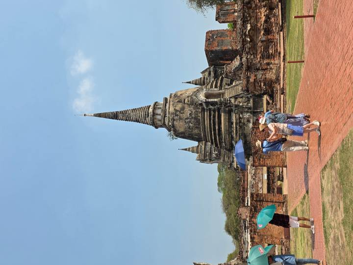 Historic brick stupa surrounded by ruined walls with tourists walking on a sunny day, some carrying colorful umbrellas