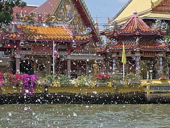 Colorful riverside shrine with ornate roofs seen through sparkling water droplets splashing up from a boat ride